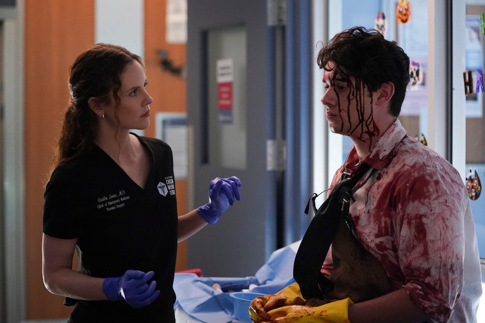 Lenox in purple gloves and black scrubs ready to treat a patient dressed as a zombie for Halloween on Chicago Med Season 10 Episode 5