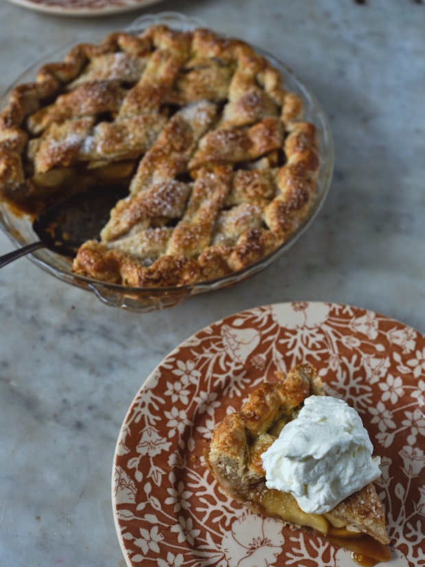 a slice of apple pie on a small plate topped with dollop of whipped cream with remaining pie in the background