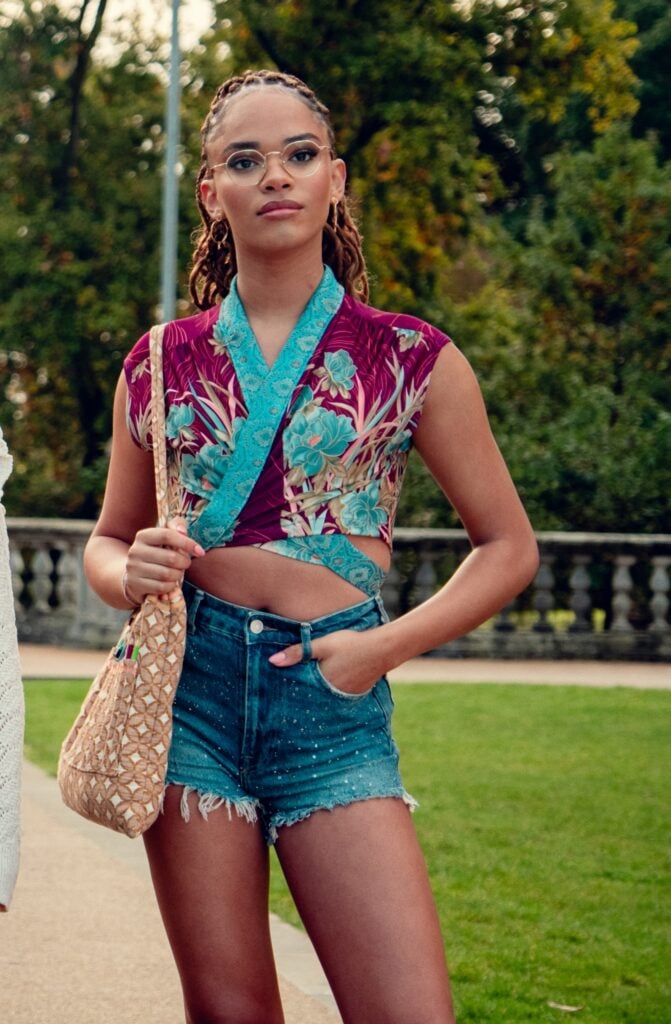 Elle in a flowered crop top with her hand in her short pocket on a beautiful college campus on Heartstopper